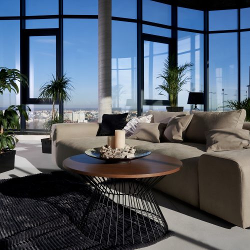 Front view of fashionable interior spacious room with large panoramic window with incredible view on city and cozy grey sofa with pillows and green flowerpots. Concept of modern interior.